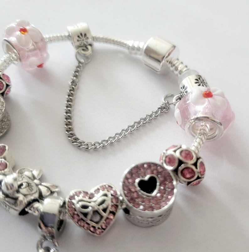 May include: A silver charm bracelet with various charms. Charms include pink flower beads, a heart-shaped charm with pink crystals, and a round charm with a black heart. The bracelet has a safety chain.