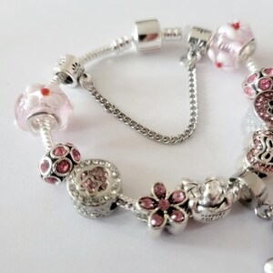 May include: A silver charm bracelet with various decorative charms. The bracelet features pink and clear glass beads with red accents, flower-shaped charms, and charms with pink gemstones. The clasp has a safety chain.