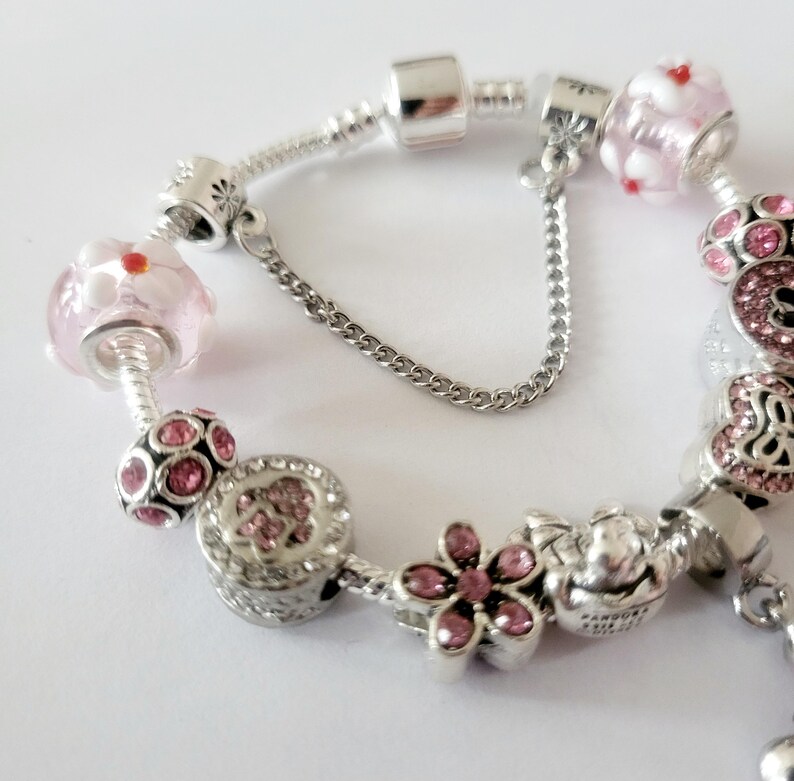 May include: A silver charm bracelet with various decorative charms. The bracelet features pink and clear glass beads with red accents, flower-shaped charms, and charms with pink gemstones. The clasp has a safety chain.