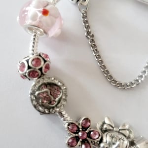 May include: A silver charm bracelet with various charms. Charms include a pink and white flower bead, a pink gem-studded bead, a charm with pink gems, a flower charm with pink gems, and a Minnie Mouse charm.