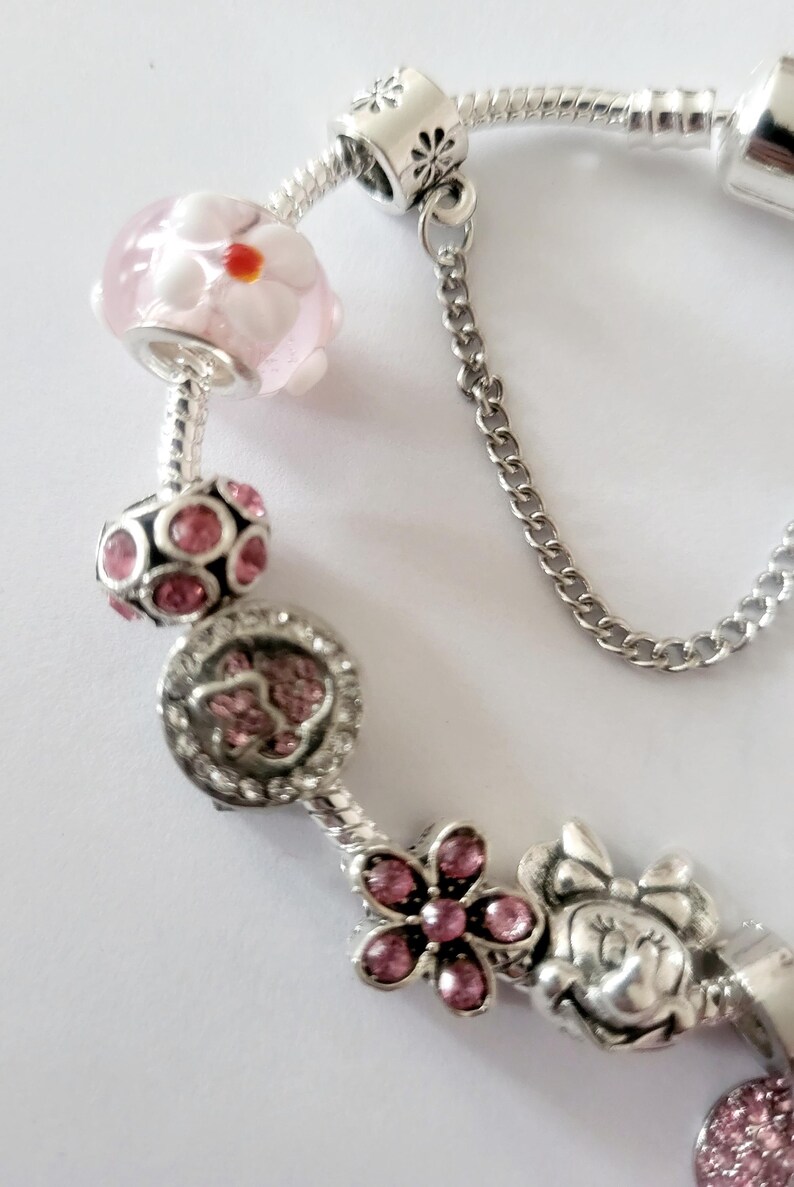 May include: A silver charm bracelet with various charms. Charms include a pink and white flower bead, a pink gem-studded bead, a charm with pink gems, a flower charm with pink gems, and a Minnie Mouse charm.
