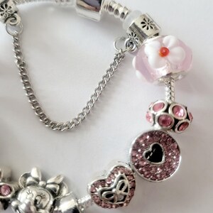 May include: A silver charm bracelet featuring pink and silver charms. The charms include a Minnie Mouse, a heart, a flower, and a round charm with a heart-shaped cutout. The bracelet has a safety chain.