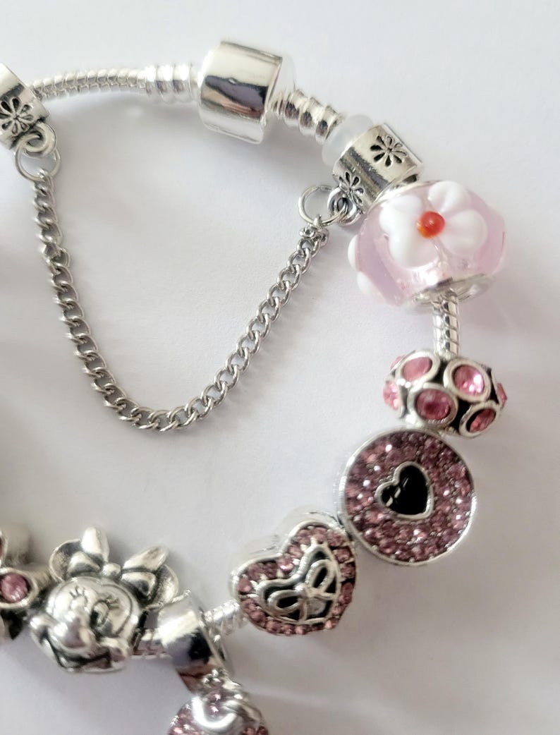 May include: A silver charm bracelet featuring pink and silver charms. The charms include a Minnie Mouse, a heart, a flower, and a round charm with a heart-shaped cutout. The bracelet has a safety chain.