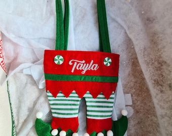 May include: Green and red elf stocking with white pom-poms and the name "Tayla" embroidered on the front.