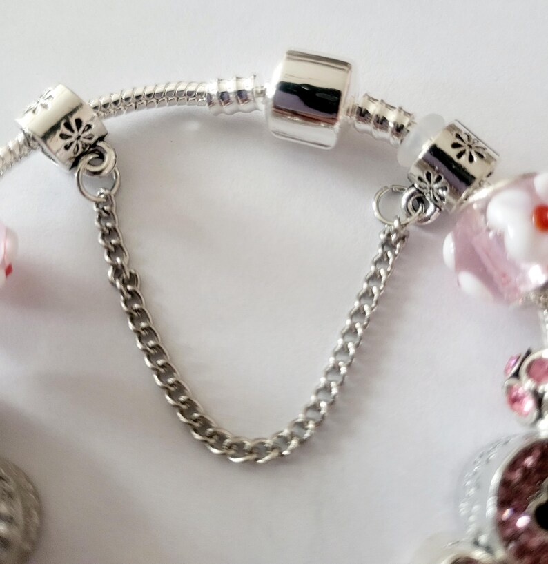 May include: Silver charm bracelet with a safety chain. The bracelet features decorative beads with floral and geometric designs. The safety chain is made of small, linked silver chain.