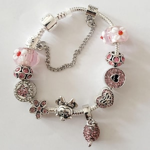 May include: A silver charm bracelet featuring pink and clear beads. The bracelet includes a Minnie Mouse charm, a flower charm, and a dangling pink charm. The beads have floral and heart designs.