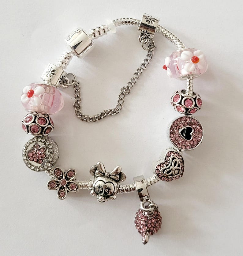 May include: A silver charm bracelet featuring pink and clear beads. The bracelet includes a Minnie Mouse charm, a flower charm, and a dangling pink charm. The beads have floral and heart designs.