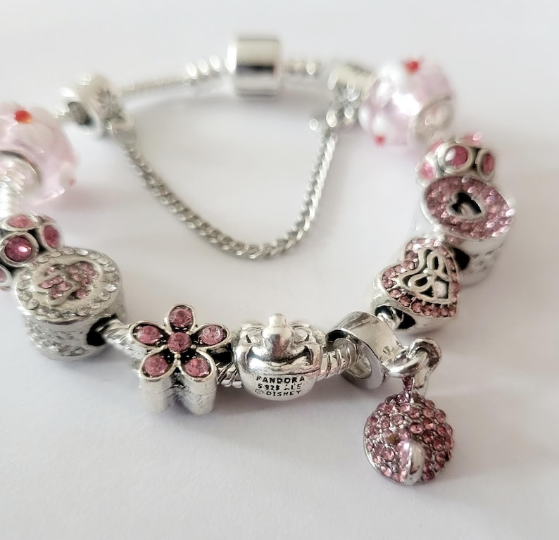 May include: A silver charm bracelet with various pink and silver charms. Charms include a flower, heart, and a dangling pink charm. The bracelet has a safety chain and several pink and silver beads.