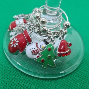 May include: A clear wine glass with silver charms. Charms include a red mitten with a white snowflake, a snowman, a green Christmas tree, and a red Santa hat. The glass sits on a green surface.
