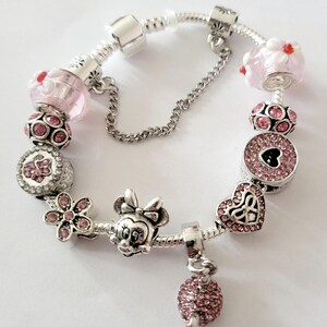 May include: A silver charm bracelet with various pink and silver charms. Charms include a Minnie Mouse head, a flower, a heart, and a round charm with a heart-shaped cutout. The bracelet has a safety chain.