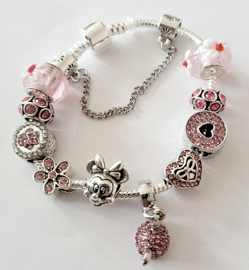 May include: A silver charm bracelet with various pink and silver charms. Charms include a Minnie Mouse head, a flower, a heart, and a round charm with a heart-shaped cutout. The bracelet has a safety chain.
