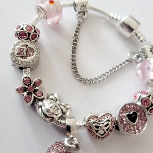 May include: A silver charm bracelet featuring pink and clear beads and charms. Charms include a flower, heart, and a round charm with a black heart. The bracelet has a safety chain.