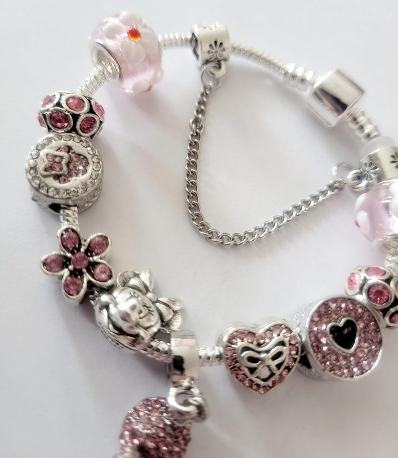 May include: A silver charm bracelet featuring pink and clear beads and charms. Charms include a flower, heart, and a round charm with a black heart. The bracelet has a safety chain.
