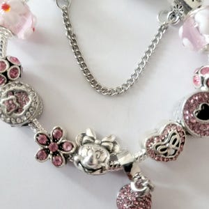 May include: A silver charm bracelet featuring pink and silver charms. Charms include a Minnie Mouse head, flowers, hearts, and a pink beaded charm. The bracelet has a safety chain.