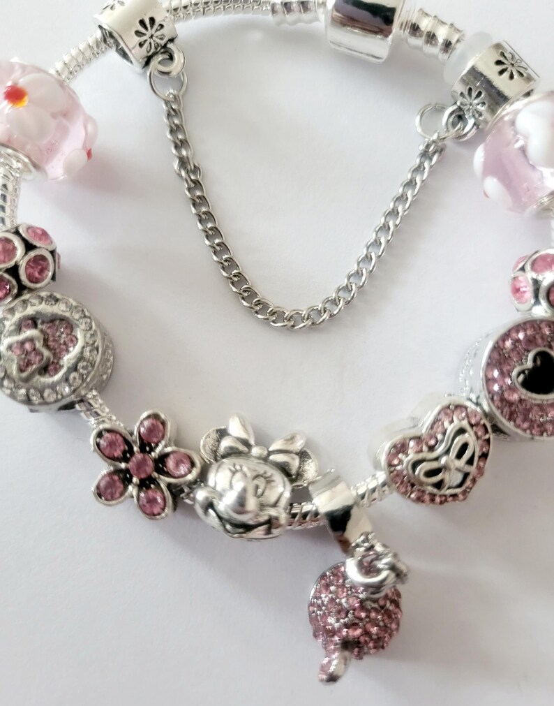 May include: A silver charm bracelet featuring pink and silver charms. Charms include a Minnie Mouse head, flowers, hearts, and a pink beaded charm. The bracelet has a safety chain.