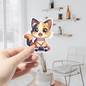 May include: A colorful sticker featuring a cartoon cat with orange, brown, and white fur, holding a video game controller. The cat has large, expressive eyes and a happy expression. The sticker is held in a person's hand.