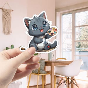May include: A cute cartoon gray cat holding a paintbrush and a colorful paint palette. The cat is sitting on a piece of paper with a paintbrush on the ground.