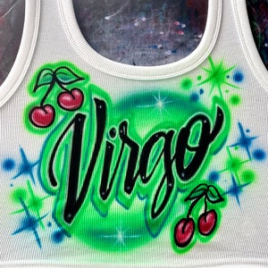 May include: White tank top with a green and blue airbrushed design featuring the word "Virgo" in black cursive lettering and two red cherries on each side.