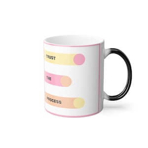 Op de afbeelding: Een witte keramische mok met een zwarte handgreep. De mok heeft een roze en geel ontwerp met de tekst "TRUST THE PROCESS" erop gedrukt.
