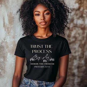 Op de afbeelding: Zwart T-shirt met de tekst "TRUST THE PROCESS" boven een bergafbeelding en de tekst "HONOR THE PROMISE PROVERBS 3:5-6". Het shirt wordt gedragen door een persoon met krullend donker haar en een spijkerbroek.