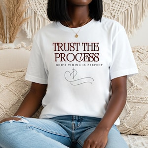 May include: White t-shirt with the text "TRUST THE PROCESS" in a dark red font. Below the text is "GOD'S TIMING IS PERFECT" and a heart-shaped design. The shirt is short-sleeved and worn with blue jeans.