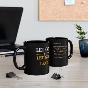 May include: Two black ceramic mugs featuring white and yellow text. One mug displays the phrase LET GO LET GOD LEAD with a small cross. The other mug features the text COMMIT YOUR WAY TO THE LORD; TRUST IN HIM AND HE WILL ACT. PSALM 37:5.