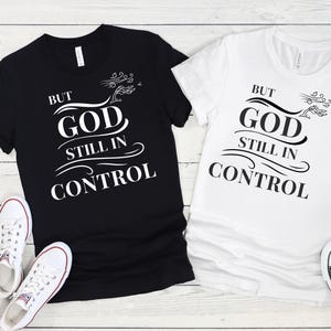 May include: Two t-shirts, one black and one white, with the text "BUT GOD STILL IN CONTROL" in a bold, stylized font. The shirts have a crew neck and short sleeves. White sneakers are visible near the black t-shirt.