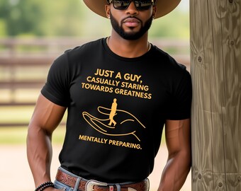 Puede incluir: Una camiseta negra con el texto "JUST A GUY, CASUALLY STARING TOWARDS GREATNESS MENTALLY PREPARING." se usa con un sombrero de vaquero marrón, gafas de sol y un cinturón marrón con hebilla plateada. La persona lleva vaqueros azules y un reloj plateado.