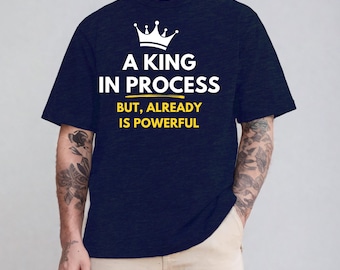 May include: Navy blue t-shirt with the text "A KING IN PROCESS" and a crown graphic. The text "BUT, ALREADY IS POWERFUL" is below the main text. The shirt is a dark blue color.