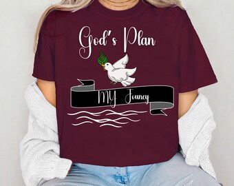 May include: A maroon t-shirt featuring the text "God's Plan My Journey" in white script. A white dove with a green sprig is above a black banner. The shirt is a casual wear item.