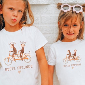 Könnte beinhalten: Zwei Kinder tragen weiße T-Shirts mit einem Motiv von zwei Affen, die ein Tandem fahren. Die Shirts sagen "Beste Freunde". Die Mädchen tragen Sonnenbrillen und haben ihre Haare zu Zöpfen gebunden.