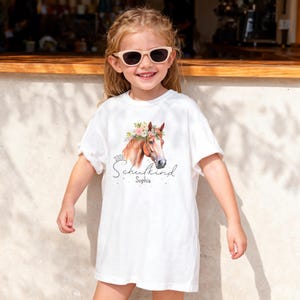 Op de afbeelding: Een wit t-shirt met een afbeelding van een bruin paard met een bloemenkrans. De tekst "2026" staat boven het woord "Schulkind" in een cursief lettertype, met de naam "Sophia" eronder in een kleiner, eenvoudig lettertype.