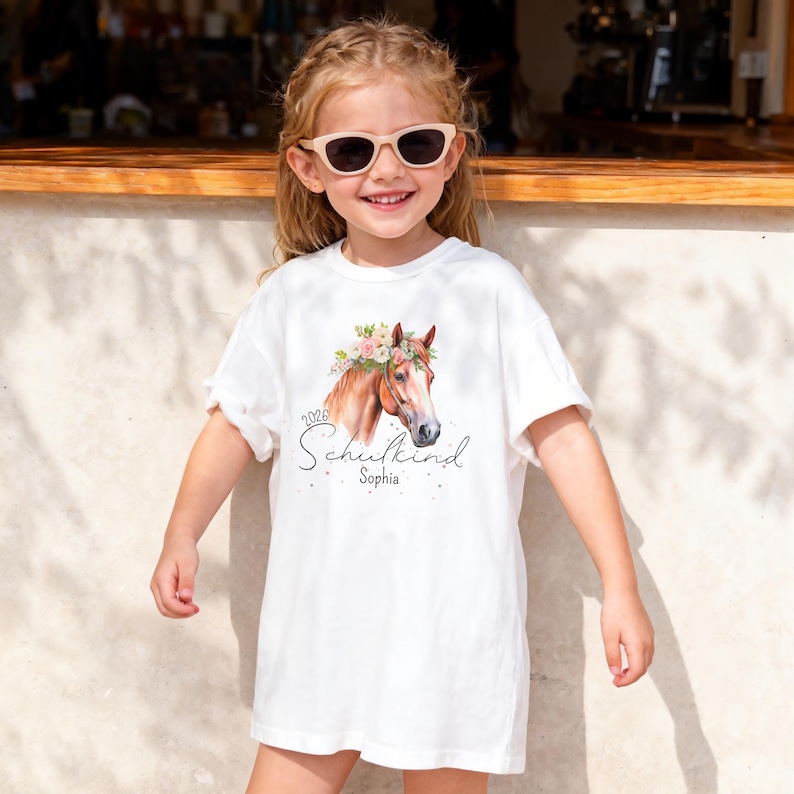 Op de afbeelding: Een wit t-shirt met een afbeelding van een bruin paard met een bloemenkrans. De tekst "2026" staat boven het woord "Schulkind" in een cursief lettertype, met de naam "Sophia" eronder in een kleiner, eenvoudig lettertype.