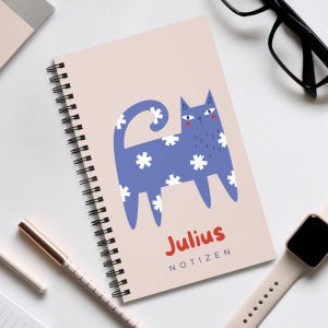 Könnte beinhalten: Ein rosa Spiralnotizbuch mit einer blauen Katzendarstellung. Die Katze hat weiße Sterne auf ihrem Körper und einen eingerollten Schwanz. Das Notizbuch trägt die Aufschrift "Julius Notizen" in roten Buchstaben.