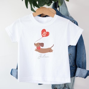 Könnte beinhalten: Weißes T-Shirt mit einem braunen Dackel, der einen roten herzförmigen Ballon mit der Zahl 5 hält. Der Hund trägt ein rotes Halstuch. Der Name "Julian" steht darunter in Schreibschrift.