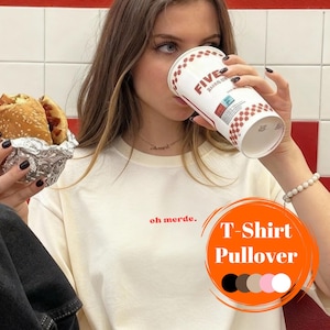 Könnte beinhalten: Cremefarbenes T-Shirt mit dem Schriftzug "oh merde." in Rot. Das Shirt ist Teil einer T-Shirt/Pullover-Kollektion, mit Farbauswahl unten. Eine Person hält einen Becher und einen verpackten Burger.