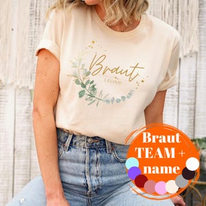 Könnte beinhalten: Ein weißes T-Shirt mit dem Text "Braut Leonie" in goldener Schrift und einem grünen floralen Kranzdesign. Das Shirt wird von einer Person mit blauer Jeans getragen.