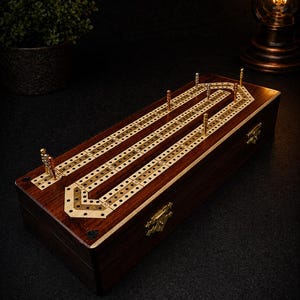 Può includere: Una tavola da cribbage rettangolare in legno con finitura marrone scuro e un inserto di colore chiaro che presenta tre piste parallele di fori. La tavola include chiusure metalliche dorate sul lato e diversi pioli metallici inseriti.