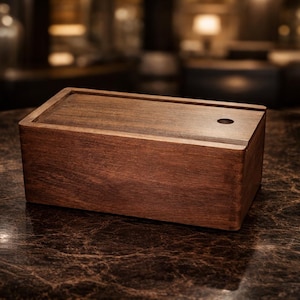 Puede incluir: Una caja rectangular de madera con tapa, con un acabado marrón oscuro. La caja tiene un pequeño agujero circular en la parte superior. Se encuentra sobre una superficie oscura y texturizada, lo que sugiere un objeto decorativo o de almacenamiento.