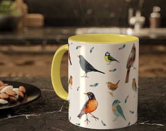 May include: A yellow and white ceramic mug with a pattern of colorful birds and feathers. The mug has a yellow rim and handle.