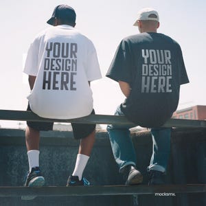 May include: Two people wearing oversized t-shirts with the text "YOUR DESIGN HERE" printed on the back. One shirt is white, the other is dark gray. They are sitting on a metal bar, with a clear sky in the background.