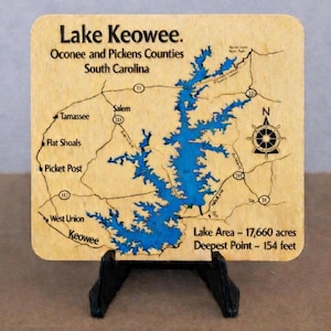 May include: A square wooden map of Lake Keowee in Oconee and Pickens Counties, South Carolina, featuring a blue resin inlay for the water, black printed text, road lines, a compass rose, and data stating Lake Area - 17,660 acres and Deepest Point - 154 feet.