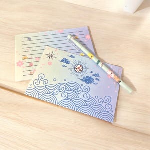 May include: Two blue and white stationery note cards with a pen. The top card has a pink and white floral design with lines for writing. The bottom card has a blue and white wave design. The pen is green and white with a black tip.