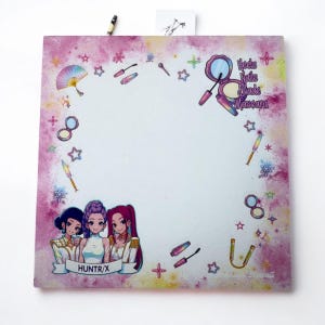 Op de afbeelding: Een vierkant, wit notitieblok met een roze en paarse aquarel-achtige rand. Het notitieblok bevat illustraties van make-up items, sterren en drie vrouwen in anime-stijl. De tekst op het notitieblok luidt "Heels Nails Blade Mascara" en "HUNTR/X".