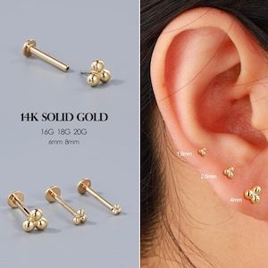 Könnte beinhalten: Goldfarbene Ohrringe aus massivem 14K Gold. Die Ohrringe haben eine Anordnung aus drei kleinen Kugeln. Das Bild enthält Text mit Größenangaben: 16G, 18G, 20G, 6 mm und 8 mm. Die Ohrringe sind auf grauem Hintergrund und an einem Ohr abgebildet.