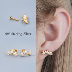 May include: Gold-toned earrings with white gemstone accents. One earring is a flat-backed stud, and the other is a curved bar with multiple gemstones. The text "925 Sterling Sliver" is visible.