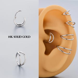 Puede incluir: Una colección de pendientes de oro macizo de 14 quilates. La imagen muestra varios estilos, incluyendo un diseño de doble aro, un aro con bisagras y múltiples estilos de puños para la oreja. Los pendientes se muestran sobre un fondo neutro y una oreja modelo.