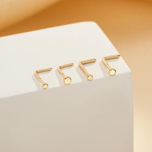 May include: Four gold-colored L-shaped nose studs with a flat, circular end. The studs are arranged in a row on a white surface, with a soft, warm-toned background.
