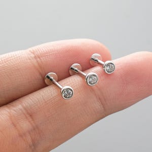 May include: Three silver-colored, flat-backed stud earrings with a small, round, clear gemstone set in a bezel. The earrings are displayed on a light-colored background.
