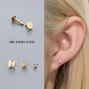 May include: Gold-colored, disc-shaped earrings with a sunburst design. The image shows multiple sizes of the earrings, along with one in a person's ear. The text "14K SOLID GOLD" is displayed. The earrings are small and delicate.
